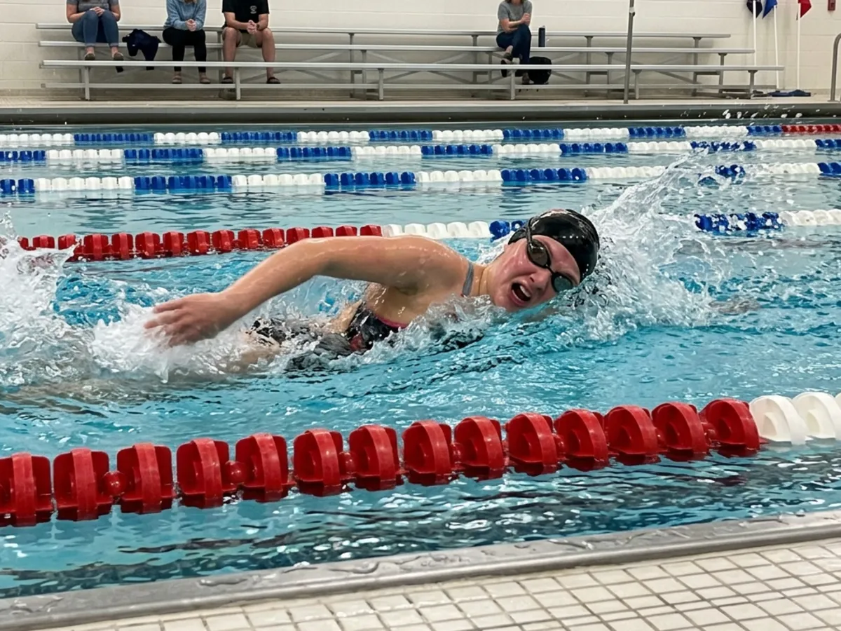 A teenage swimmer mid-stroke during a small-town swim meet, parents watching from the bleachers.